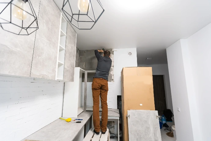 Carpenter standing on a step stool using a power drill to install upper wall cabinets during a modern kitchen remodeling project.