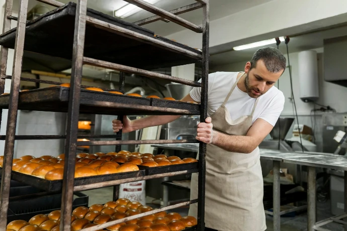 Baker in an apron moving a commercial rolling rack filled with freshly baked golden bread buns in a bakery.