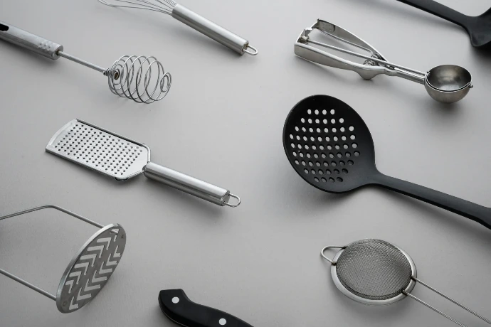 A flat lay collection of stainless steel and black plastic kitchen utensils, including a whisk, grater, potato masher, and skimmer spoon on a grey surface.