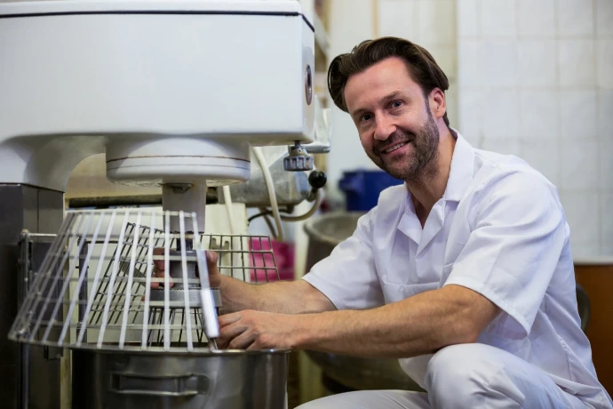 A smiling professional baker in white uniform operating a large commercial dough mixer in a bakery kitchen.