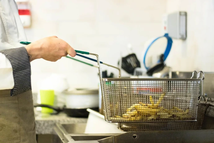 A chef lifting a stainless steel deep fryer basket filled with freshly cooked french fries in a restaurant kitchen.