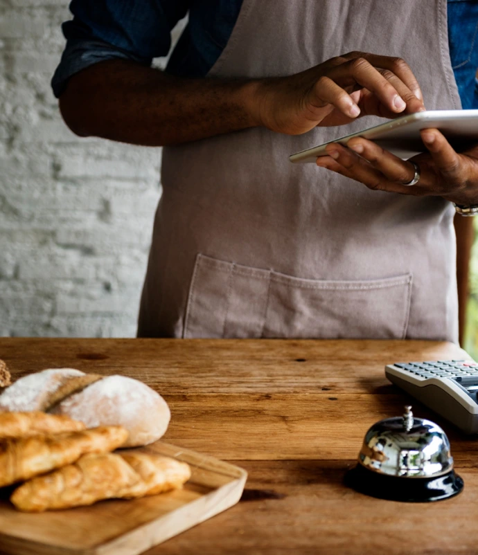 A bakery worker using a digital tablet at a checkout counter displaying fresh croissants, bread, and bagels next to a POS terminal.